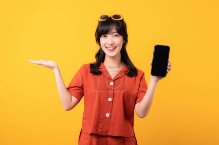 Téléchargez les photos : Portrait jeune belle femme asiatique sourire heureux habillé de vêtements orange avec smartphone et geste de la main pointant vers l'espace libre isolé sur fond de studio jaune. app smartphone concept - en image libre de droit