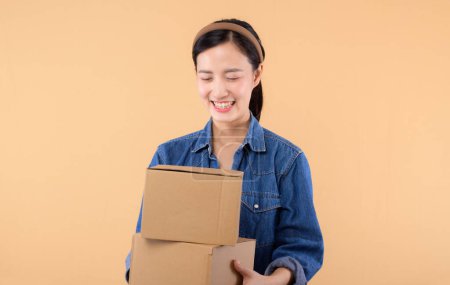 Foto de Retrato de mujer asiática joven con camisa de mezclilla que sostiene la caja de paquete aislado en fondo beige estudio, mensajero de entrega y concepto de servicio de envío. - Imagen libre de derechos