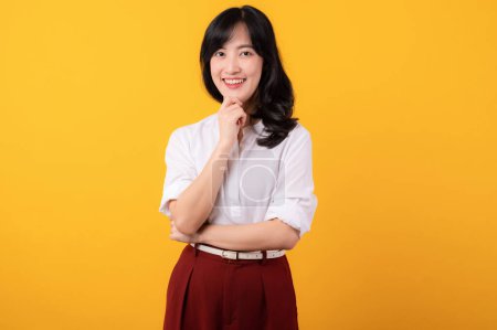 Foto de Retrato hermosa joven asiática mujer emprendedor feliz sonrisa vestida con camisa blanca y plantas rojas mostrando pensamiento gesto aislado en amarillo estudio fondo. - Imagen libre de derechos