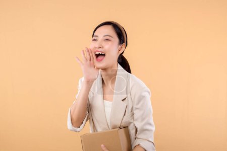 Téléchargez les photos : Portrait de heureux heureux confiant jeune femme d'affaires asiatique portant une veste blanche tenant boîte de colis isolé sur fond de studio beige, livraison messager et concept de service d'expédition. - en image libre de droit