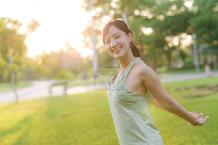 Téléchargez les photos : Joggeuse. Adaptez-vous à une jeune femme asiatique avec des vêtements de sport verts respirant de l'air frais dans le parc avant de courir et de profiter d'un extérieur sain. Fitness fille coureuse dans le parc public. Concept de bien-être - en image libre de droit