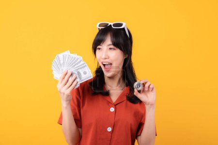 Téléchargez les photos : Portrait belle jeune femme asiatique sourire heureux habillé en vêtements orange tenant de l'argent comptant et crypto Bitcoin monnaie numérique célébrer son succès isolé sur fond jaune. Être riche concept. - en image libre de droit