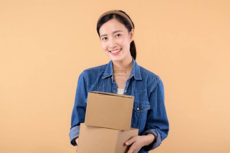 Téléchargez les photos : Portrait jeune femme asiatique portant chemise en denim tenant boîte de colis isolé sur fond de studio beige, livraison messager et concept de service d'expédition. - en image libre de droit