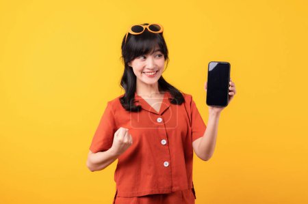 Téléchargez les photos : Portrait jeune belle femme asiatique heureux sourire habillé en vêtements orange avec smartphone et poing geste de la main isolé sur fond de studio jaune. app smartphone concept - en image libre de droit