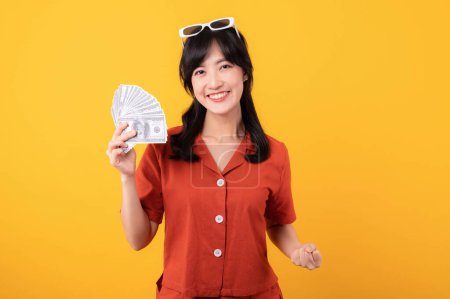 Foto de Retrato hermosa joven asiática mujer feliz sonrisa vestida con ropa naranja sosteniendo dinero en efectivo y celebrar su éxito aislado sobre fondo amarillo. Ser rico concepto. - Imagen libre de derechos