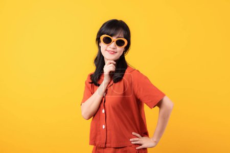 Téléchargez les photos : Portrait belle jeune femme asiatique sourire heureux habillé en vêtements orange et lunettes de soleil regardant célébrer avec la main sur son visage isolé sur fond de studio jaune. - en image libre de droit