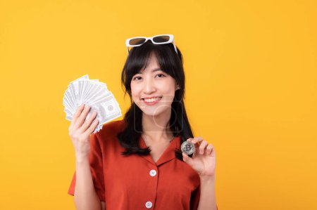 Photo for Portrait beautiful young asian woman happy smile dressed in orange clothes holding cash money and crypto bitcoin digital currency celebrate her success isolated on yellow background. Be rich concept. - Royalty Free Image