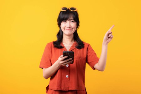 Téléchargez les photos : Portrait jeune belle femme asiatique heureux sourire vêtu de vêtements orange avec smartphone et pointant doigt geste de la main pour libérer de l'espace isolé sur fond de studio jaune. app smartphone concept - en image libre de droit