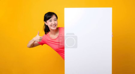 Téléchargez les photos : Asiatique jeune sport fitness femme heureux sourire portant des vêtements de sport rose debout derrière la bannière blanche vierge ou vide espace panneau publicitaire sur fond jaune. bien-être concept de style de vie. - en image libre de droit
