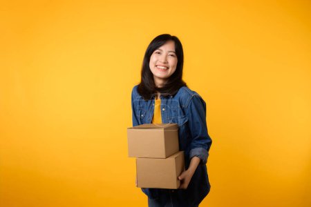 Photo for Portrait young asian woman wearing yellow t-shirt and denim shirt holding parcel box isolated on yellow studio background, Delivery courier and shipping service concept. - Royalty Free Image