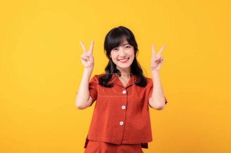 Téléchargez les photos : Portrait belle jeune femme asiatique sourire heureux habillé en vêtements orange montrant deux doigts signe de victoire geste de main isolé sur fond de studio jaune. - en image libre de droit
