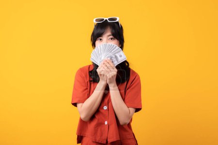 Foto de Retrato hermosa joven asiática mujer feliz sonrisa vestida con ropa naranja mostrando dinero en efectivo aislado sobre fondo amarillo. Pagar y comprar concepto de pago de compras. - Imagen libre de derechos