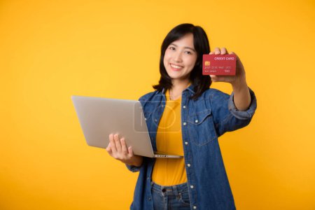 Téléchargez les photos : Jeune femme heureuse d'origine asiatique porter t-shirt jaune chemise en denim en utilisant ordinateur portable pc détenir carte bancaire de crédit achats livraison de commande en ligne isolé sur fond jaune uni. - en image libre de droit