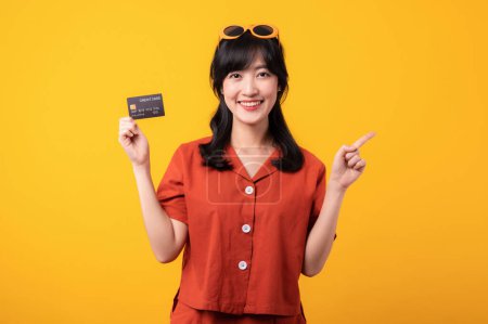 Téléchargez les photos : Portrait belle jeune femme asiatique sourire heureux habillé de vêtements orange montrant carte de crédit et pointeur vers l'espace libre isolé sur fond jaune. Paiement et achat shopping concept de paiement. - en image libre de droit