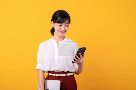 Foto de Retrato joven hermosa mujer asiática emprendedor feliz sonrisa usando camisa blanca y plantas rojas usando teléfono inteligente y la celebración de aislamiento portátil sobre fondo amarillo. - Imagen libre de derechos