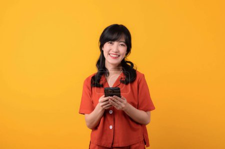 Photo for Portrait young beautiful asian woman happy smile dressed in orange clothes using a smartphone isolated on yellow studio background. app smartphone concept - Royalty Free Image