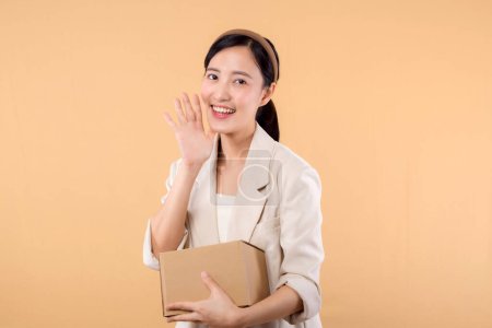 Téléchargez les photos : Portrait de heureux heureux confiant jeune femme d'affaires asiatique portant une veste blanche tenant boîte de colis isolé sur fond de studio beige, livraison messager et concept de service d'expédition. - en image libre de droit