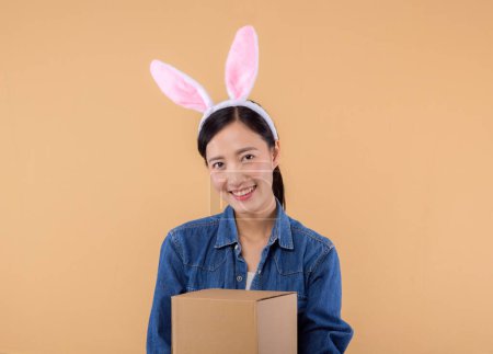 Foto de Retrato de mujer asiática joven con orejas de conejo conejo y camisa de mezclilla que sostiene la caja de paquete aislado en fondo beige estudio, mensajero de entrega y concepto de servicio de envío. - Imagen libre de derechos