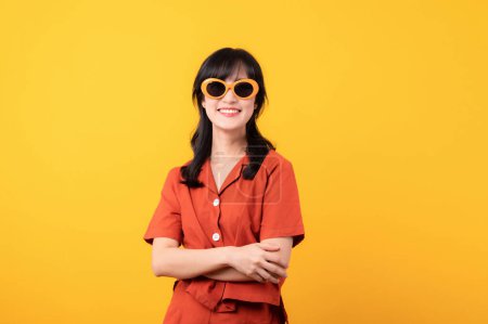 Foto de Retrato hermosa joven asiática mujer feliz sonrisa vestida con ropa naranja y gafas de sol cruzando brazos expresar confianza emocional sensación aislada sobre fondo amarillo. - Imagen libre de derechos