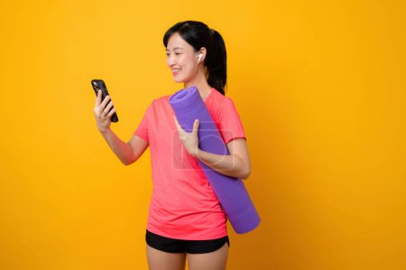Foto de Retrato asiático joven deportista fitness mujer sonrisa feliz usando ropa deportiva rosa y teléfono inteligente haciendo ejercicio de entrenamiento con fondo de estudio amarillo. tecnología bienestar estilo de vida concepto. - Imagen libre de derechos