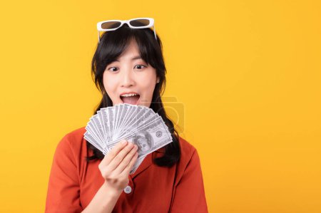 Foto de Retrato hermosa joven asiática mujer feliz sonrisa vestida con ropa naranja mostrando dinero en efectivo aislado sobre fondo amarillo. Pagar y comprar concepto de pago de compras. - Imagen libre de derechos