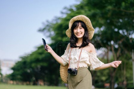 Photo for Portrait of young asian woman traveler with weaving hat, basket, mobile phone and camera on green public park background. Journey trip lifestyle, world travel explorer or Asia summer tourism concept. - Royalty Free Image