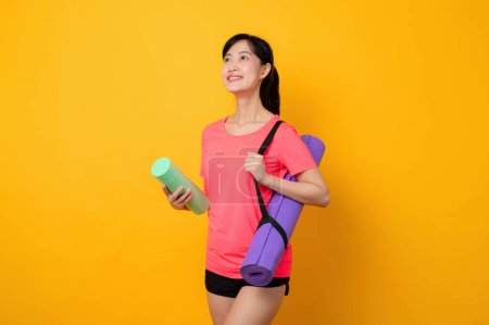 Téléchargez les photos : Portrait asiatique jeune sport fitness femme heureux sourire portant des vêtements de sport rose et tapis de yoga faire de l'exercice séance d'entraînement sur fond jaune studio. Concept de mode de vie sain. - en image libre de droit