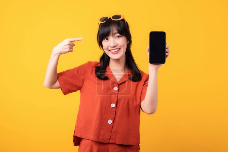 Téléchargez les photos : Portrait jeune belle femme asiatique sourire heureux habillé de vêtements orange pointant doigt geste de la main au smartphone isolé sur fond de studio jaune. app smartphone concept - en image libre de droit