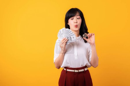 Photo for Portrait young beautiful asian woman enterpriser with doubt face wearing white shirt and red plants holding cash dollar money and crypto digital currency isolation on yellow background. Wealth concept - Royalty Free Image