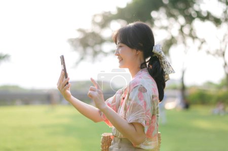 Photo for Portrait of young asian woman traveler with weaving basket, mobile phone on green public park background. Journey trip lifestyle, world travel explorer or Asia summer tourism concept. - Royalty Free Image