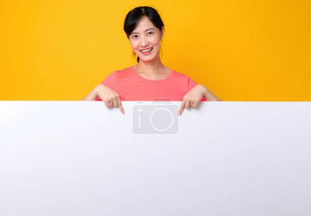 Foto de Asiático joven deportes fitness mujer feliz sonrisa usando rosa ropa deportiva de pie detrás de la bandera blanca en blanco o espacio vacío tablero de anuncios sobre fondo amarillo. bienestar concepto de estilo de vida. - Imagen libre de derechos