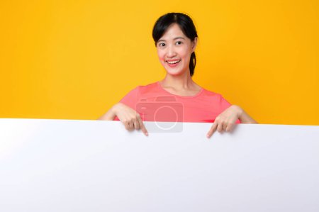 Photo for Asian young sports fitness woman happy smile wearing pink sportswear standing behind the white blank banner or empty space advertisement board against yellow background. wellbeing lifestyle concept. - Royalty Free Image
