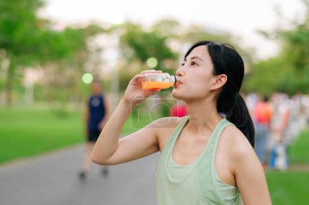 Photo for Female jogger. Fit Asian young woman with green sportswear drinking organic orange juice after running and enjoying a healthy outdoor. Fitness runner girl in public park. Wellness being concept - Royalty Free Image