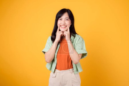Foto de Mujer asiática alegre de 30 años con camisa verde y naranja. Con los dedos apuntando a los dientes, enfatiza la salud dental y la sonrisa radiante aislada en el fondo amarillo. promover el concepto de cuidado oral. - Imagen libre de derechos