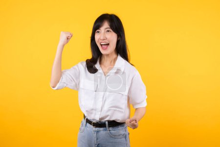 Téléchargez les photos : Portrait belle asiatique jeune femme sourire heureux avec poing vers le haut expression geste de victoire joyeuse sa réussite sur fond de studio jaune. Journée femme gagnant concept de célébration. - en image libre de droit