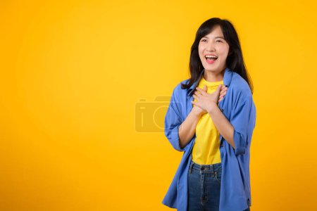 Erleben Sie die Wärme und Freude mit einem herzerwärmenden Porträt. Eine junge Asiatin in blauem Hemd zeigt ein glückliches Lächeln, während sie ihre Hand auf der Brust hält. echtes Glück und ein Gefühl der Dankbarkeit.