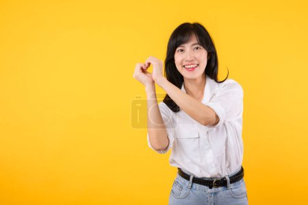 Photo for Young healthcare female. Asian woman wearing white shirt feels happy and romantic shapes heart gesture expresses tender feeling poses isolated on yellow background. People affection and care concept. - Royalty Free Image