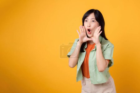 Foto de Mujer asiática alegre de 30 años vistiendo camisa naranja y camisa verde mostrando gesto de mano cubriendo la boca, simbolizando grito o sorpresa. Momentos energéticos y sentido de exuberancia. - Imagen libre de derechos