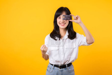 Téléchargez les photos : Jeune femme asiatique tenant une carte de crédit devant un œil et montrant un geste de poing à la main avec sourire heureux isolé sur fond jaune. Paiement achats concept en ligne. - en image libre de droit