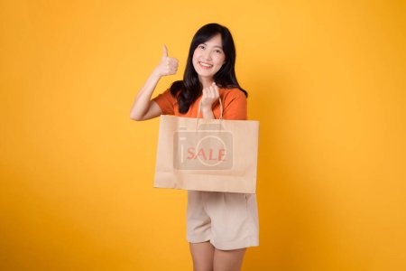 Téléchargez les photos : Portrait heureuse jeune femme asiatique trentenaire séduisante avec robe printanière tendance, chemise orange mode et sac en papier isolé sur fond jaune. Vente d'été concept de shopping. - en image libre de droit