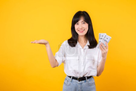 Téléchargez les photos : Une jeune femme heureuse d'origine asiatique porte une chemise blanche et un jean en jean tenant de l'argent comptant en dollar et pointant la main pour libérer de l'espace sur fond jaune. Investissement et concept financier. - en image libre de droit