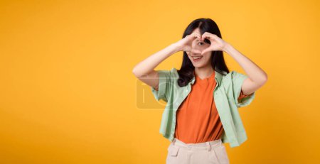 Téléchargez les photos : Jeune femme asiatique trentenaire portant une chemise verte et orange. Avec un geste de main de coeur sur l'œil, ajoute une touche fantaisiste. Expression unique et créative d'affection sur un fond jaune vif. - en image libre de droit