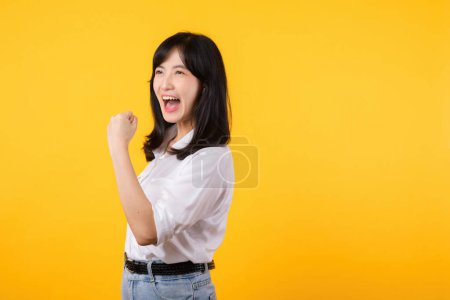 Téléchargez les photos : Portrait belle asiatique jeune femme sourire heureux avec poing vers le haut expression geste de victoire joyeuse sa réussite sur fond de studio jaune. Journée femme gagnant concept de célébration. - en image libre de droit