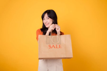 Photo for Portrait happy attractive young 30s asian woman with trendy springtime dress, orange shirt fashion and paper bag isolated on yellow background. Summertime sale shopping concept. - Royalty Free Image