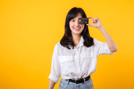 Foto de Mujer asiática joven sosteniendo la tarjeta de crédito delante de un ojo con sonrisa feliz aislado en el fondo amarillo. Compras de pago concepto online. - Imagen libre de derechos