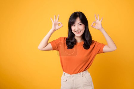 Photo for Radiating positivity, Asian cheerful woman 30s wearing orange shirt shows okay hand gesture against yellow background, symbolizing approval and assurance. Positivity, agreement, confidence concept. - Royalty Free Image
