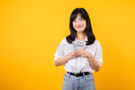 Téléchargez les photos : Heureuse jeune femme d'affaires asiatique portant une chemise blanche et un jean en jean tenant de l'argent, embrassant l'argent des dollars et souriant, debout sur fond jaune. Amour d'être riche concept. - en image libre de droit