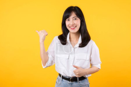 Photo for Happy asian young woman wearing white shirt and denim jean plants pointing thumb finger away on copy space isolated on yellow background. helps pick best choice, recommends product concept. - Royalty Free Image