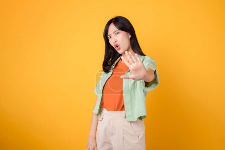 Foto de Mujer asiática alegre de 30 años con camisa verde y naranja. Con una mano levantada en gesto de 'no', expresa convicción aislada sobre fondo amarillo. - Imagen libre de derechos