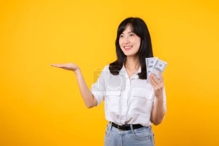 Téléchargez les photos : Une jeune femme heureuse d'origine asiatique porte une chemise blanche et un jean en jean tenant de l'argent comptant en dollar et pointant la main pour libérer de l'espace sur fond jaune. Investissement et concept financier. - en image libre de droit